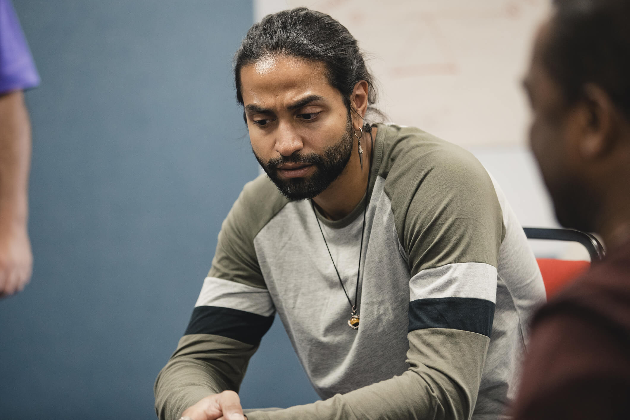 A man with long hair sits at a table, engaged in conversation with another man.