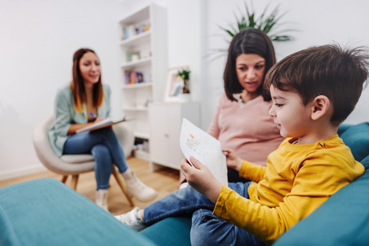 A young boy holds a drawing as his mother and therapist watch him