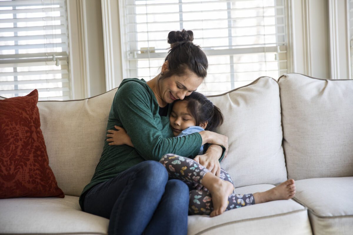 In a living room, a mother hugs her young daughter as they sit on a white sofa